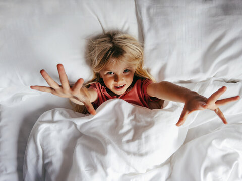 Child girl waking up in a bed at home, lying under blanket in cozy bedroom with white bedding set, family lifestyle childhood morning routine kid stretching and relaxing candid emotions