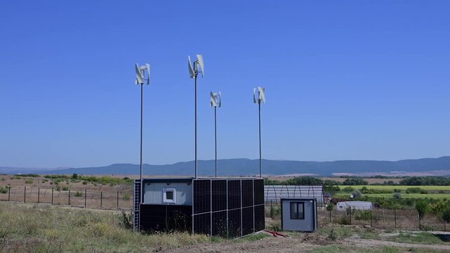 A small, off-grid container building clad entirely in solar panels sits in a rural field alongside four spinning vertical wind turbines under a bright, clear blue sky.