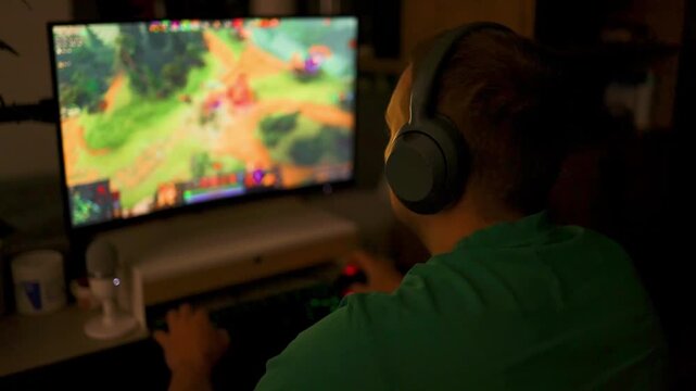Over the shoulder view of a boy wearing headphones playing a colorful multiplayer strategy or shooter game on a monitor in a dark room.