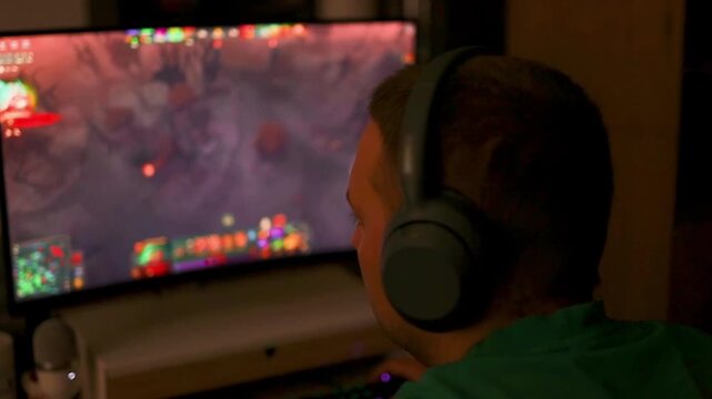 Over the shoulder view of a boy wearing headphones playing a colorful multiplayer strategy or shooter game on a monitor in a dark room.