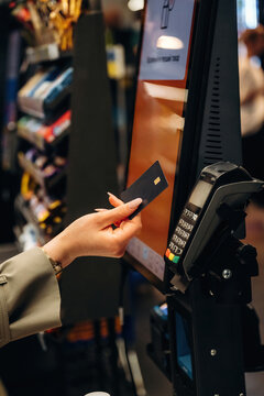 Hand of a woman holding a credit card near a contactless payment terminal at a retail checkout with various products visible in the background