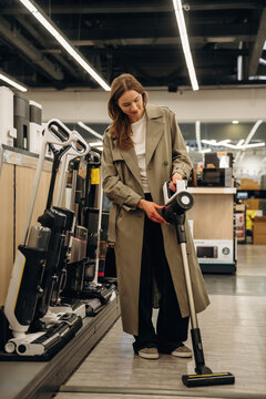 Woman in a long beige trench coat examines a cordless vacuum cleaner in a home appliance store aisle with various vacuum models displayed on shelves