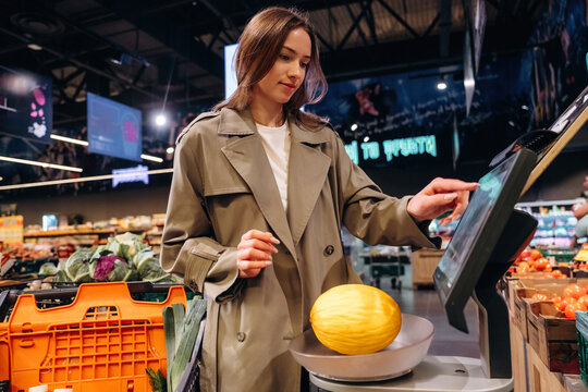 Female shopper in beige trench coat weighing yellow melon on digital scale in modern grocery store with colorful produce and shopping baskets in the background