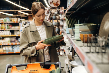Fototapeta premium Young woman shopping for kitchenware in a home goods store, examining a green plate while standing near shelves filled with various dishes and utensils