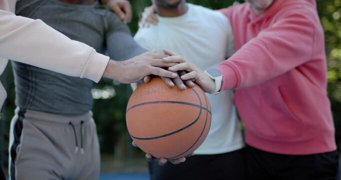 Diverse male friends holding basketball stacking hands on park court for unity under sun glare
