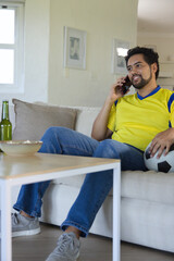 Fototapeta premium African man sitting at home wearing yellow jersey, holding soccer ball talking on phone, copy space
