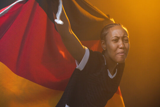 African American woman leaning waving big black-red-gold flag in stadium wearing jersey, copy space