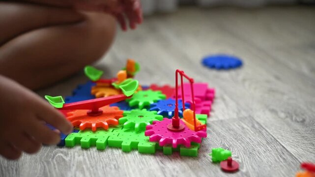 Gear toy in child hands during indoor play on wooden floor, colorful plastic gear pieces connected on base plate. educational activity mechanism supports fine motor skill. early learning concept.