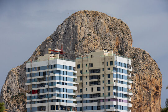 View of modern high-rise residential buildings with blue and white facades and a crane, set against the dramatic limestone cliffs of the Penon de Ifach in Calp, Valencian Community, Spain.