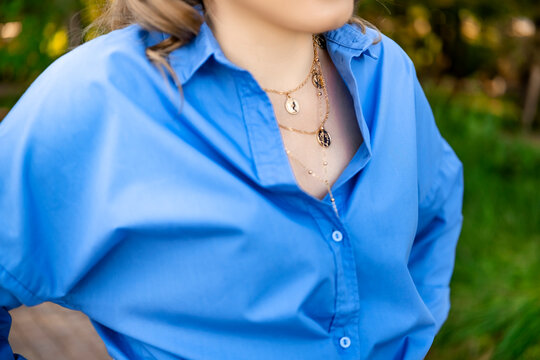 Woman wearing layered gold necklace with coin pendants and unbuttoned blue shirt outdoors. Modern jewelry trend and elegant summer outfit.