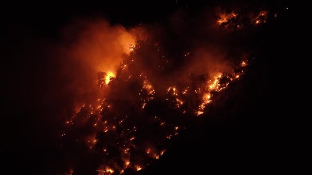 Aerial night footage of a devastating forest fire raging down a steep mountain slope with bright flames and dense smoke.