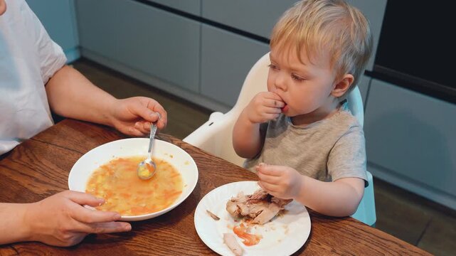 Blond toddler eating from small plate while adult feeds spoonful beside wooden kitchen table in calm home dining scene with side view, soft light, modern cabinetry, food texture
