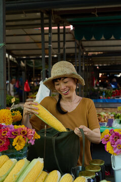 Asian Woman Shopping For Fresh Corn At A Colorful Farmers Market Stall Wearing Straw Hat