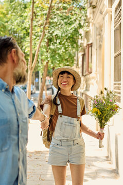 Asian Woman Smiling With Bouquet Holding Hands With Partner On Sunny Urban Street
