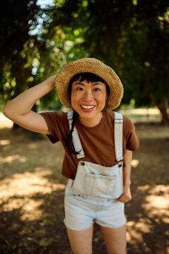 Smiling Asian Woman in Straw Hat and Denim Overalls Posing in Sunny Park for Lifestyle Branding
