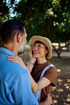 Couple Embracing in Park With Asian Woman in Straw Hat Smiling and Touching Partner's Face