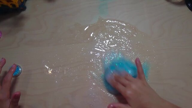 A top-down overhead shot shows a preteen child kneading glossy blue glitter slime made from PVA glue and activator across a smooth wooden desk during a hands-on chemistry science craft project.
