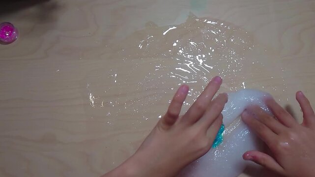 A top-down overhead shot shows a preteen child reaching for a tub of blue glitter while kneading a clear PVA slime base across a wooden desk surface during a fun STEM chemistry craft project.