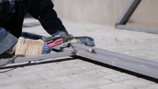 Worker bending over and working on a metal, in outdoor construction site with concrete ground, with metal structure, tools, from a close-up ground-level perspective perspective, nearby objects.