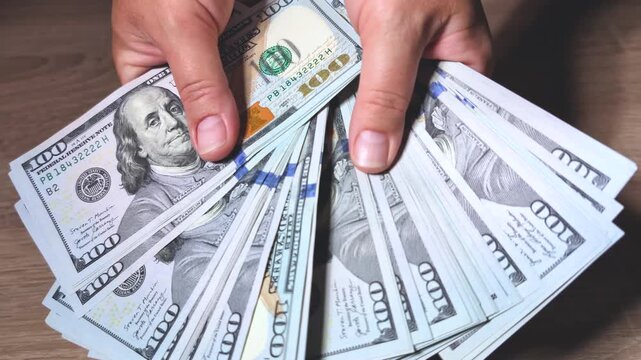 Close-up video of female hands holding and flipping through a stack of hundred dollar bills, estimating amount and enjoying feeling of financial wealth, success, and abundance. Slow motion video