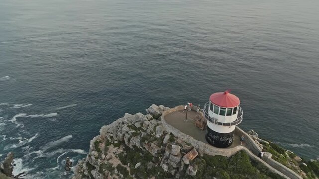 Drone view of Cape Point lighthouses perched on steep green cliffs above Atlantic waves, showing rocky ridges, narrow paths and sea foam across Cape Peninsula, South Africa in clear daylight.