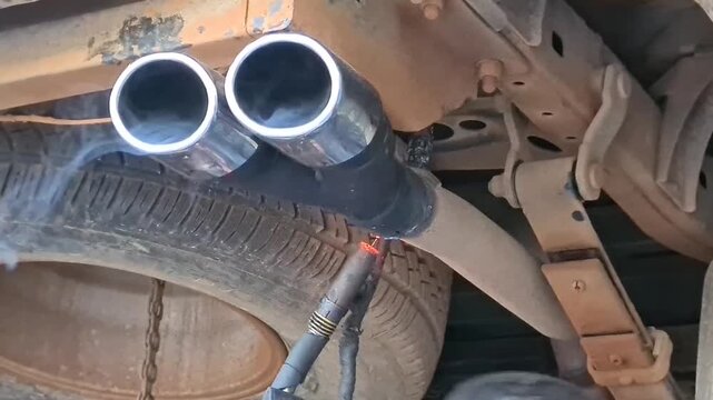 Mechanic welding a custom dual exhaust tip onto a vehicle's undercarriage. Sparks fly as the new chrome part is attached to the old exhaust pipe during auto repair.