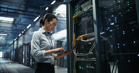 Asian Woman Network Engineer Using Laptop Computer for Server Configuration and Cable Management Beside an Open Rack Cabinet in a Big Modern Data Center Hosting Cloud Storage and AI Supercomputers.