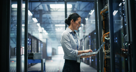 Fototapeta premium Asian Female System Administrator Working on Laptop Computer for Network Cable Management and Node Connectivity Checks Beside an Open Server Rack Cabinet With Fiber Optic Wiring in AI Data Center.