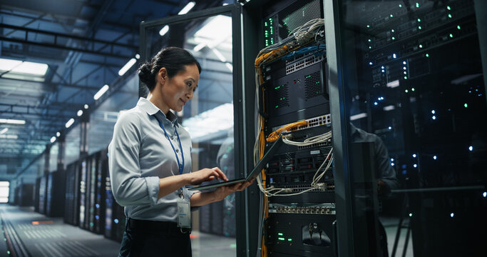 Asian Woman Network Engineer Using Laptop Computer for Server Configuration and Cable Management Beside an Open Rack Cabinet in a Big Modern Data Center Hosting Cloud Storage and AI Supercomputers.