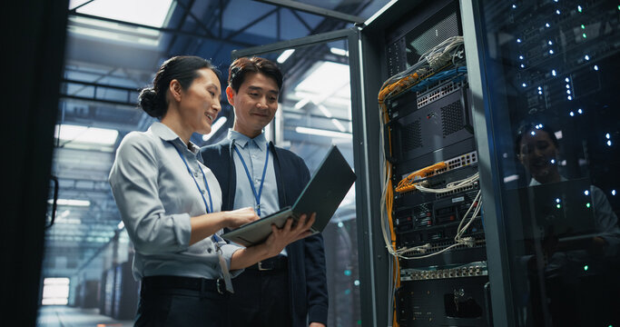 Two Asian It Engineers Talk Beside Open Server Rack Cabinet, Working on Laptop Computer for Data Backup and Network Configuration, Portrait of Male and Female Technicians in Data Center
