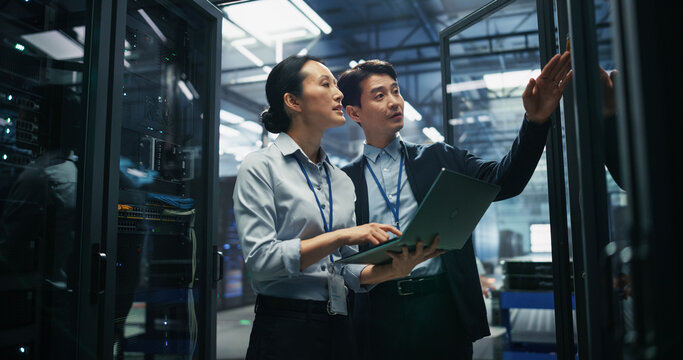 Asian Male Technical Supervisor Reaching Toward Server Rack Cabinet Door While Asian Female Database Administrator Using Laptop Computer for Equipment Inventory Audit in AI Data Center Facility.