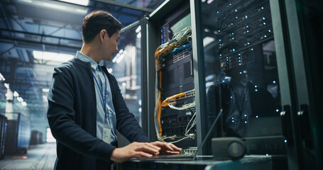 Fototapeta premium Asian Male It Support Technician With Id Badge Using Laptop Computer for Server Diagnostics and Network Troubleshooting Beside Open Rack Cabinet With Ethernet Cables in AI Data Center Facility