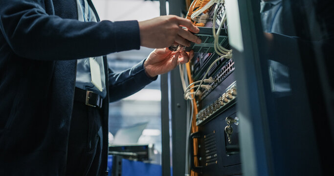 Low Angle View of Male Engineer Hands Connecting Fiber Optic Cable Into Server Rack Patch Panel, Performing Network Infrastructure Setup and Hardware Repair in Modern AI Data Center Facility.