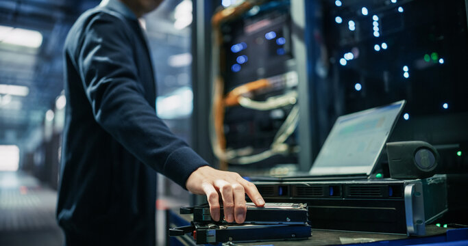 Cropped Shot of Male Data Center Technician Taking Solid State Drive From Stack on Equipment Cart While Using Laptop Computer for Storage Diagnostics Beside Rackmount Server Unit in AI Facility