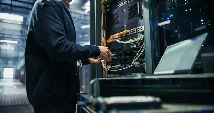 Close up of Male Data Center Engineer Hands Installing Hard Drive Into Storage Array Chassis Beside Patch Panel Cabling While Using Laptop Computer for Server Monitoring and Network Maintenance
