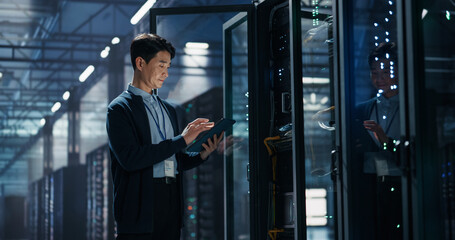 Asian Male It Specialist Using Tablet Computer to Configure Network Settings Beside Open Server Rack, Managing Data Migration and Uptime Analytics in AI Cloud Computing Colocation Facility