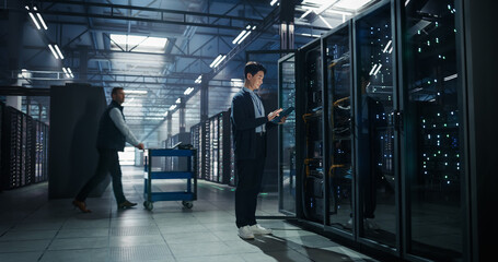 Obraz na płótnie Canvas Asian Male Systems Administrator Holding Tablet Computer at Open Server Rack While Colleague Transports Hardware on Cart, Storage Capacity and Firmware Updates in AI Cloud Computing Data Center