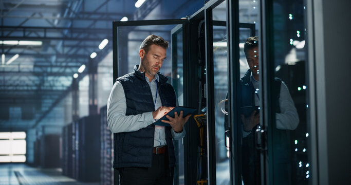 Caucasian Male Network Engineer Operating Tablet Computer at Server Rack, Running Diagnostics Software and Cybersecurity Check in AI Cloud Computing Data Center Warehouse