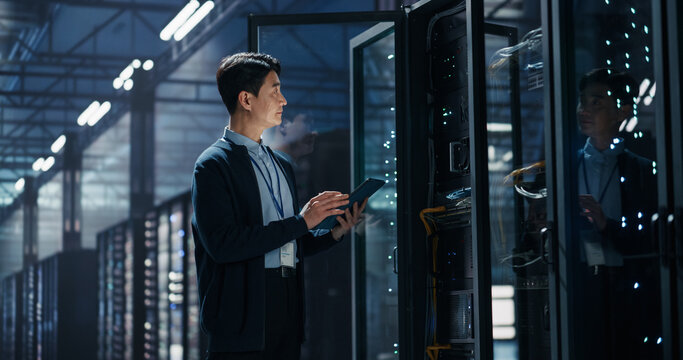 Asian Male System Administrator Holding Tablet Computer Beside Opened Server Rack Cabinet, Performing Firmware Update and Data Backup Procedure in Big Data AI Cloud Computing Data Center Facility