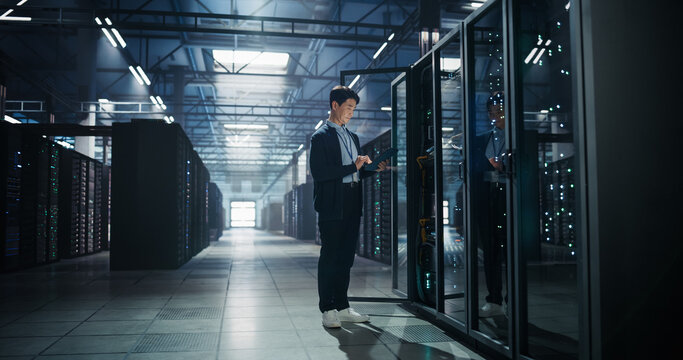 Asian Male It Specialist Standing in Aisle of Server Farm Using Tablet Computer for Network Configuration and Data Migration Tasks, Managing Machine Learning Infrastructure in AI Data Center