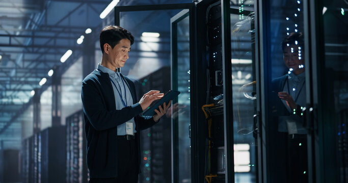 Asian Male It Specialist Using Tablet Computer to Configure Network Settings Beside Open Server Rack, Managing Data Migration and Uptime Analytics in AI Cloud Computing Colocation Facility