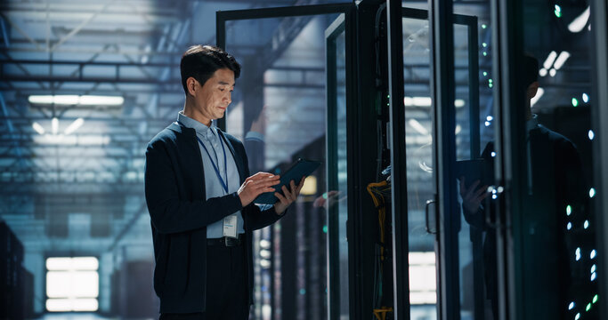 Asian Male Network Engineer With Id Badge Working on Digital Tablet Computer Next to Open Server Rack Cabinet, Running Cybersecurity Diagnostics and Bandwidth Optimization in AI Cloud Data Center