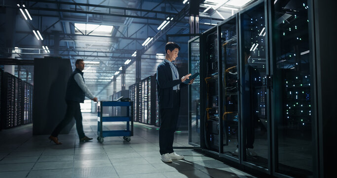 Asian Male Systems Administrator Holding Tablet Computer at Open Server Rack While Colleague Transports Hardware on Cart, Storage Capacity and Firmware Updates in AI Cloud Computing Data Center
