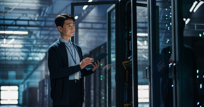 Asian Male Data Center Technician With Tablet Computer Inspecting Server Rack Hardware, Conducting Equipment Audit and Performance Monitoring in AI Cloud Computing Colocation Warehouse