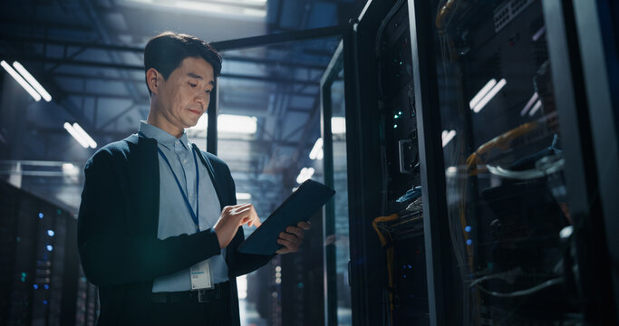 Asian Male Systems Administrator With Id Badge Working on Tablet Computer by Opened Server Rack, Conducting Firmware Updates and Bandwidth Analysis of AI Cloud Storage Hardware in Data Center