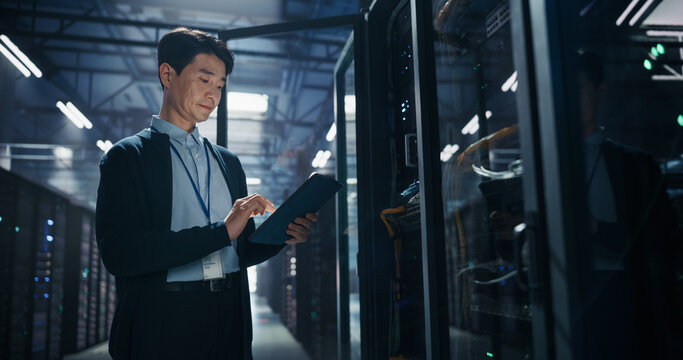 Asian Male Network Engineer Operating Tablet Computer Next to Open Server Cabinet, Running Cybersecurity Diagnostics and Database Backup Checks on AI Supercomputer Hardware in Colocation Facility