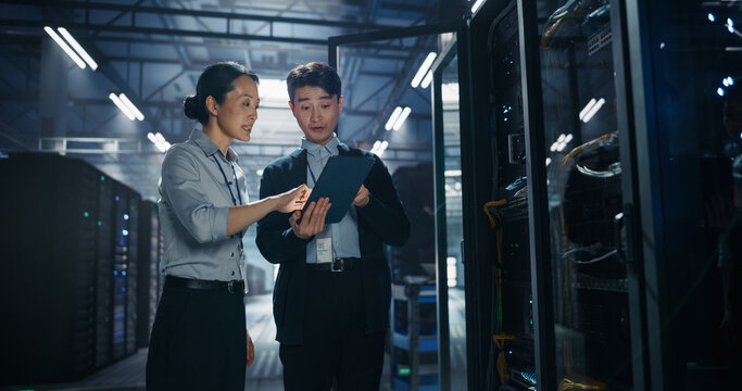 Asian Female and Male It Specialists Discussing Data on Tablet Computer by Open Server Rack, Collaborating on Network Troubleshooting and AI Server Maintenance in Big Data Center Facility
