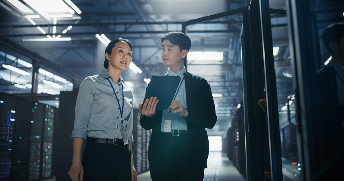 Asian Woman Looks up at Server Racks While Male Colleague Carries Tablet Computer, Two Data Center Employees Walking Through Corridor During Facility Inspection and AI Hardware Capacity Survey