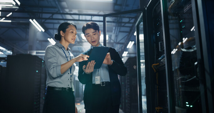 Asian Woman Points at Tablet Computer Screen While Male Colleague Holds Device, Two It Engineers Reviewing System Diagnostics and Planning AI Server Upgrade Beside Open Rack in Data Center