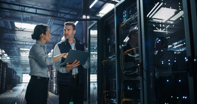 Asian Female It Specialist Talks With Caucasian Male Technician Holding Laptop Computer for Network Troubleshooting and Database Maintenance Next to Open Server Rack Cabinet in Data Center Facility